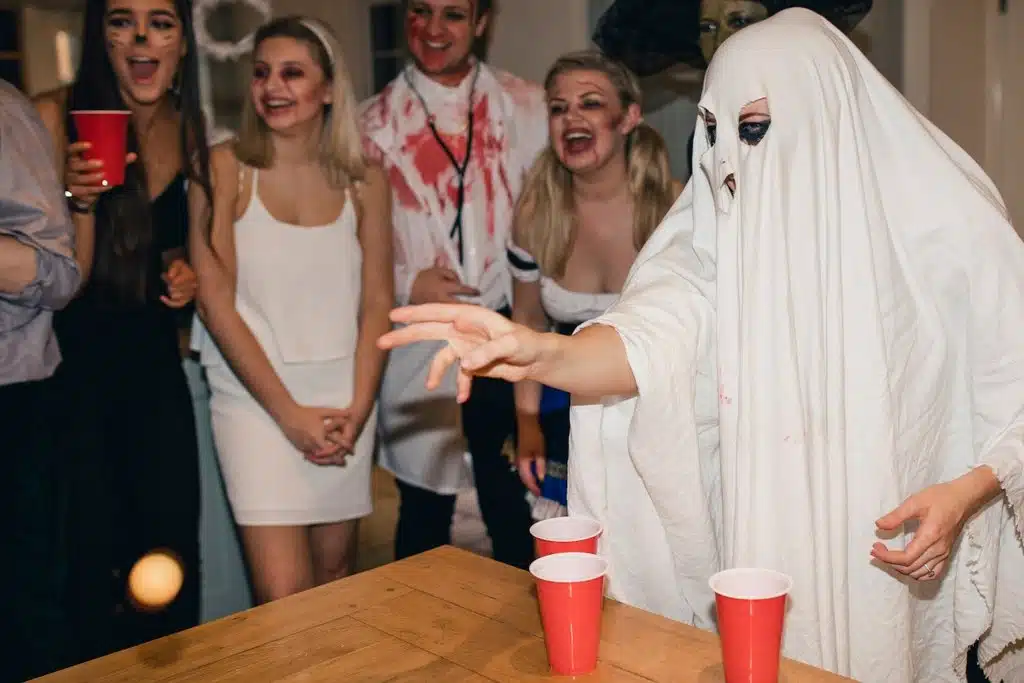 People dressed in halloween costumes. One player dressed as a ghost with a white sheet over them is throwing a pingpong ball.