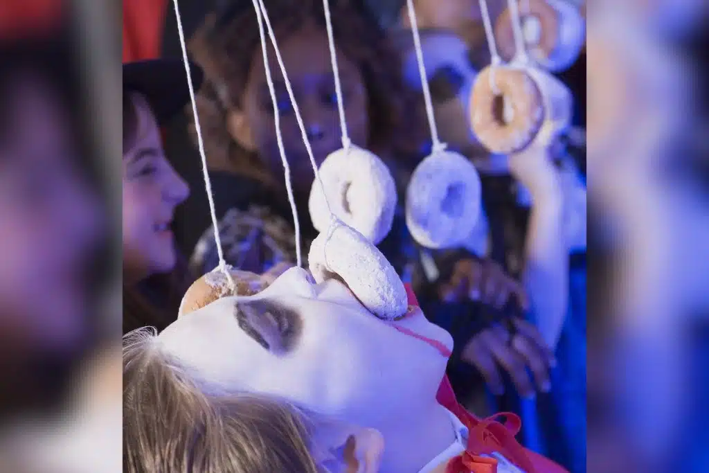 Kids dressed in Halloween costumes, trying to eat sugar dusted donuts hanging from strings.