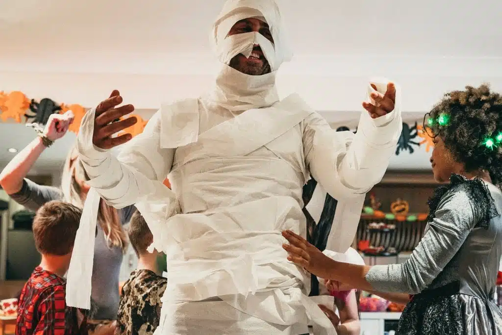 A man wrapped in tissue paper being wrapped up by a girl to look like a Halloween mummy.