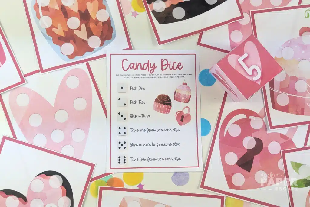 Valentine’s Day candy dice game printable with pink heart-themed game cards, a dice template, and a candy dice instruction sheet laid out on a table.